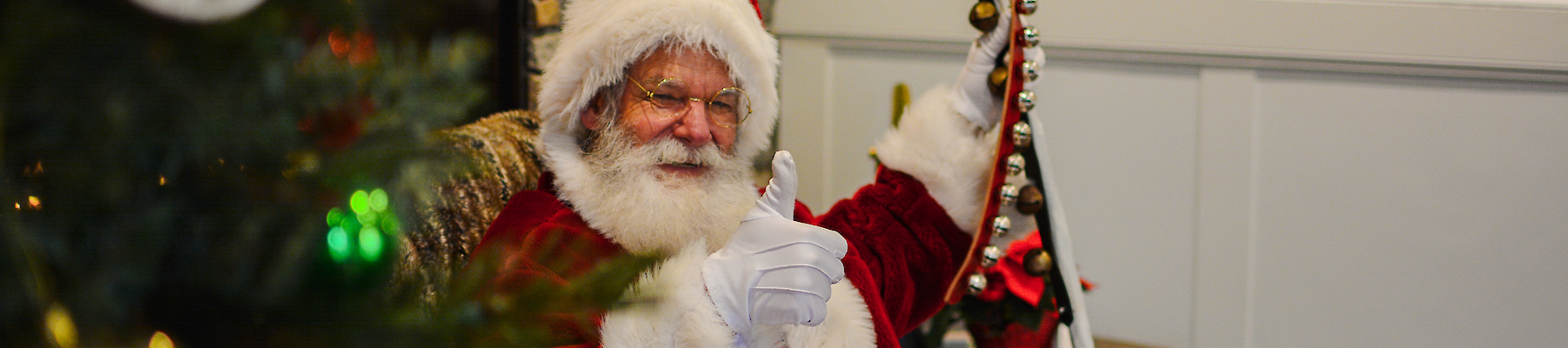 A person dressed as Santa Claus is sitting indoors, giving a thumbs-up. A decorated Christmas tree is partially visible in the foreground.