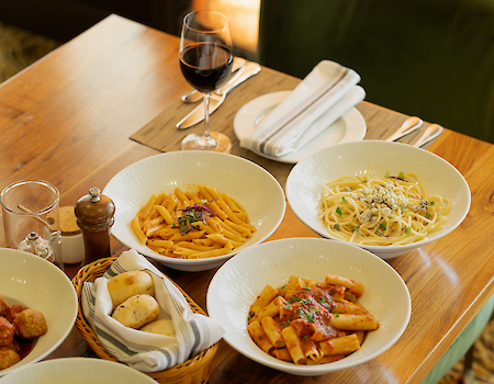 The image shows a table with pasta dishes, meatballs, bread, and a glass of red wine, set for dining.