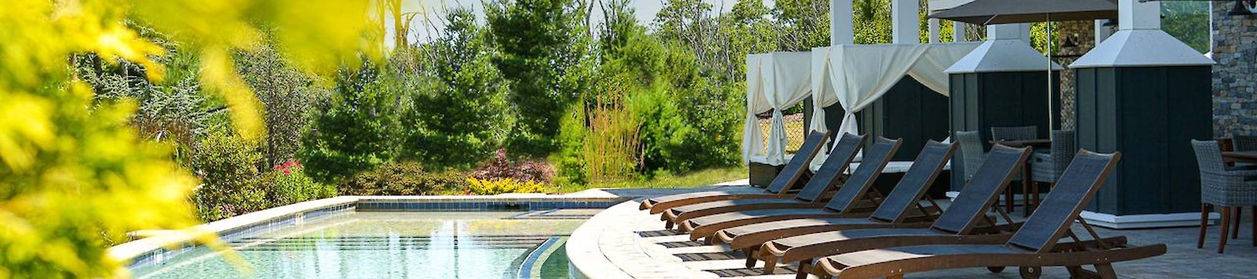 The image shows a serene poolside with lounge chairs, umbrellas, and a modern building, surrounded by greenery and open sky.