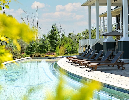 The image shows a serene poolside with lounge chairs, umbrellas, and a modern building, surrounded by greenery and open sky.