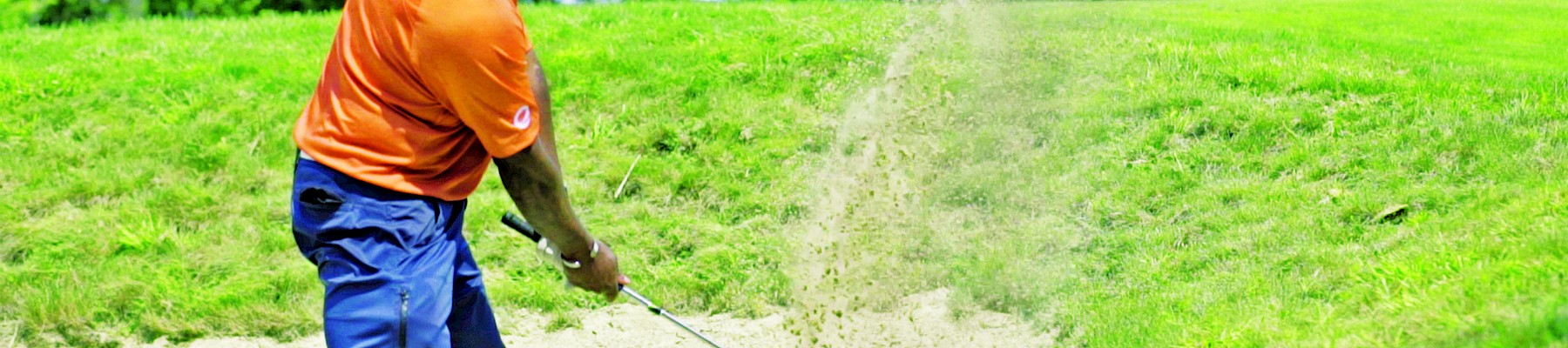 A golfer in an orange shirt plays a shot from a sand bunker, with sand flying, surrounded by lush greenery.