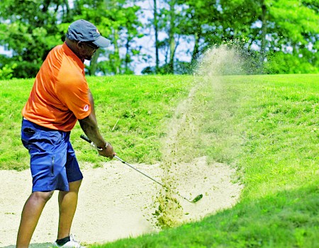 A golfer in an orange shirt plays a shot from a sand bunker, with sand flying, surrounded by lush greenery.