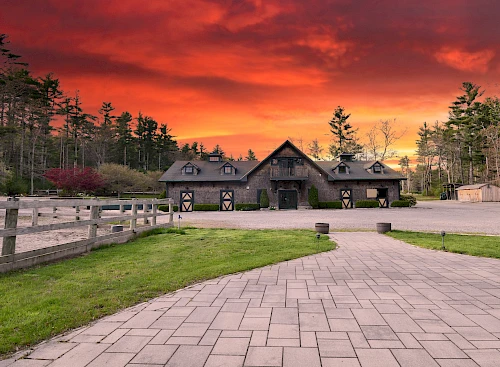 A rustic barn with a fenced paddock is under a vibrant, fiery sunset sky, surrounded by trees and a pathway leading up to it.