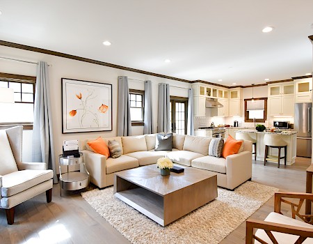 The image shows a modern living room and kitchen with a beige sectional sofa, armchairs, and a wooden coffee table on a rug.
