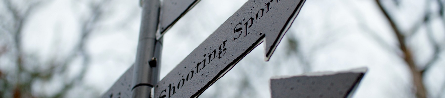 The image shows a close-up of a directional signpost with arrows pointing to "Golf", "Shooting Sports", and "Trials" against a blurred background.