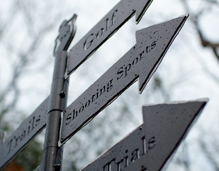 The image shows a close-up of a directional signpost with arrows pointing to "Golf", "Shooting Sports", and "Trials" against a blurred background.