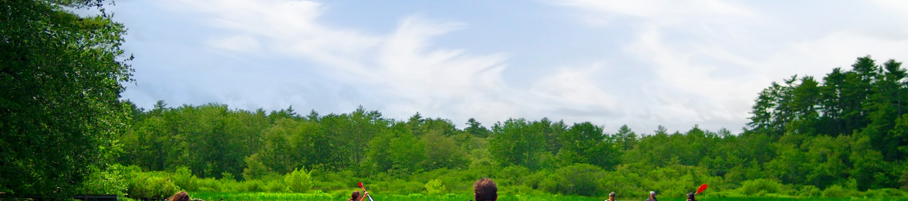 People are kayaking on a calm river surrounded by lush greenery and a clear blue sky with scattered clouds.
