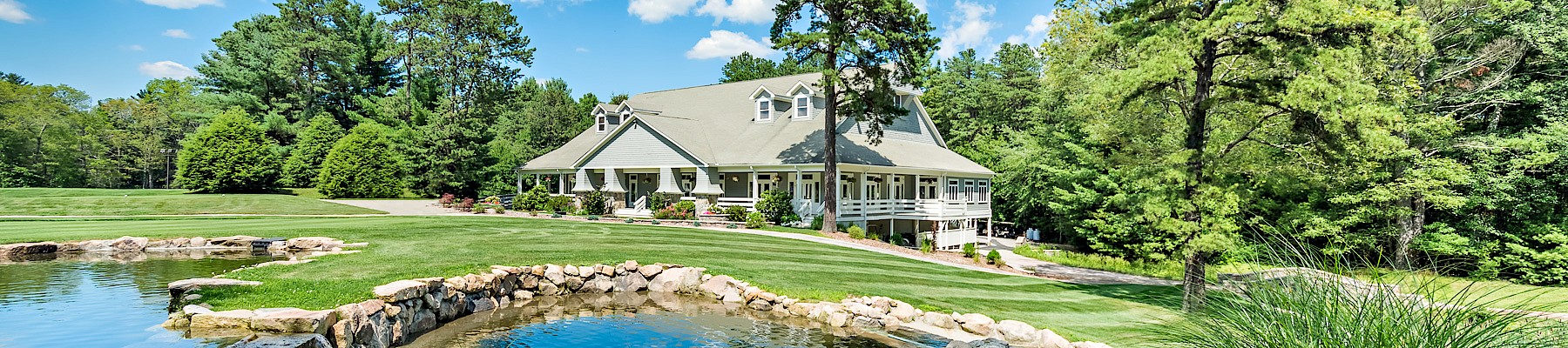 A scenic view of a large house surrounded by lush trees, a grassy lawn, and a small pond in the foreground under a bright blue sky.