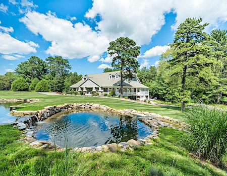 A scenic view of a large house surrounded by lush trees, a grassy lawn, and a small pond in the foreground under a bright blue sky.