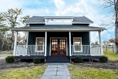 A small, charming house with a porch, surrounded by trees and a simple lawn, under a partly cloudy sky.