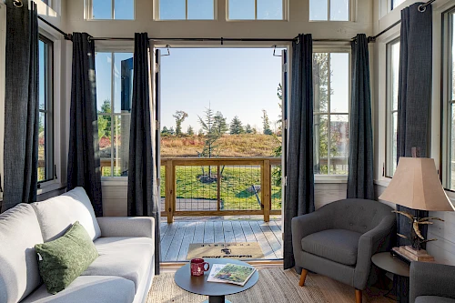 A cozy room with a sofa, armchair, and lamp, opening to a sunny deck with a view of trees and a grassy field outside.