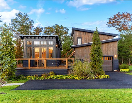 A modern, wood-paneled house with large windows, surrounded by greenery, under a clear sky, viewed from the driveway.